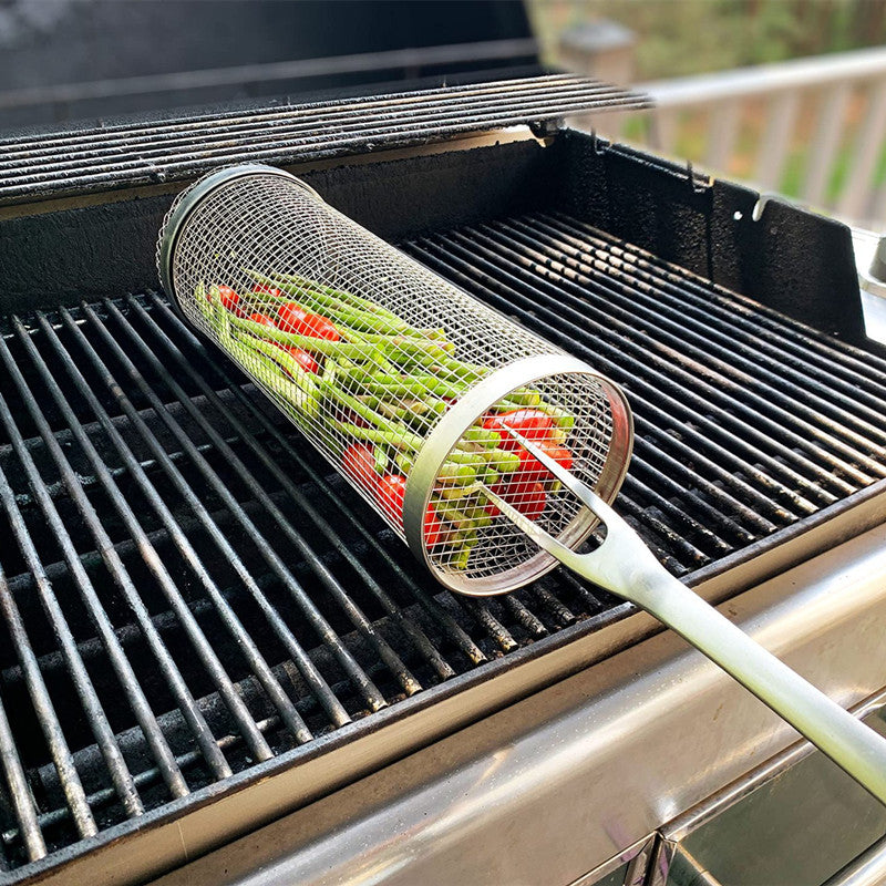 Grilling basket cylinder on grill grates with green peppers and tomatoes, long handle.